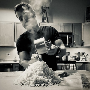 A person in a kitchen grating a large pile of steaming food, likely cheese or potatoes, onto a wooden table. The background features wooden cabinets and kitchen appliances. The image is in black and white.